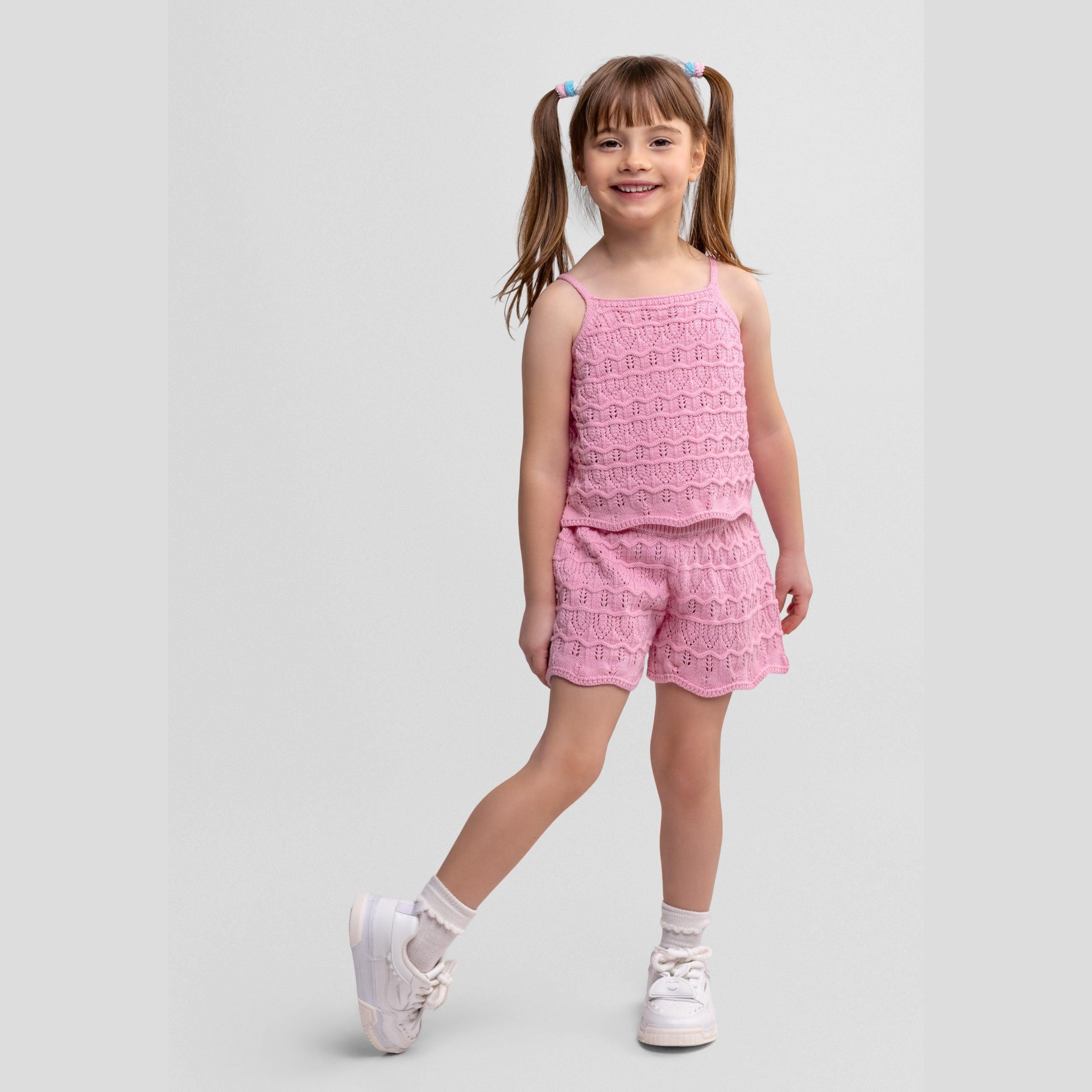 Young girl wearing pink crochet two-piece set with sleeveless top and shorts, standing against white background with pigtails and white sneakers