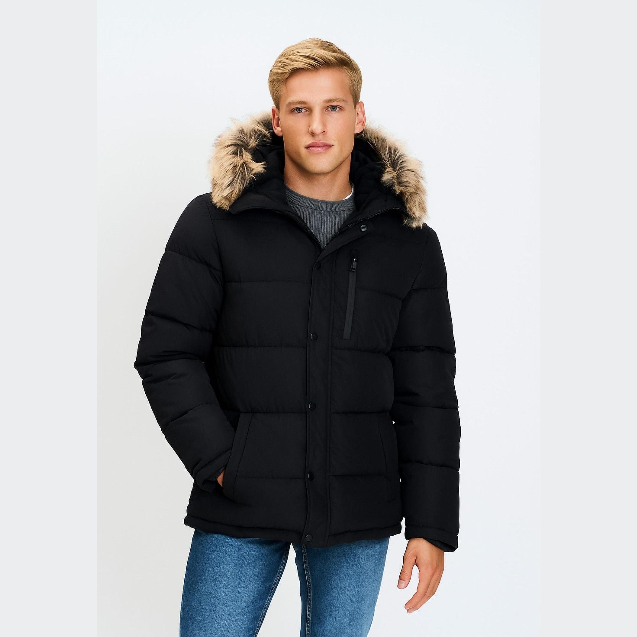 Young man wearing black padded jacket with faux fur hood trim over grey sweater and blue jeans, standing against white background