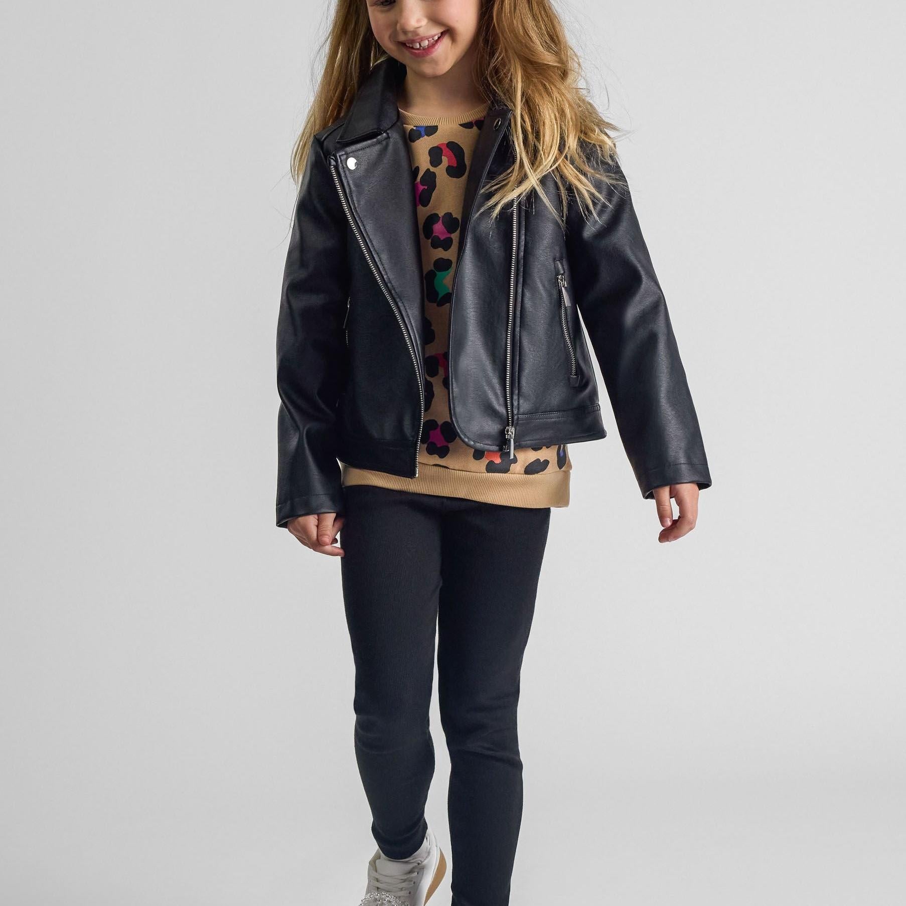 Young girl wearing black faux leather biker jacket over leopard print top with dark jeans, smiling against white studio background