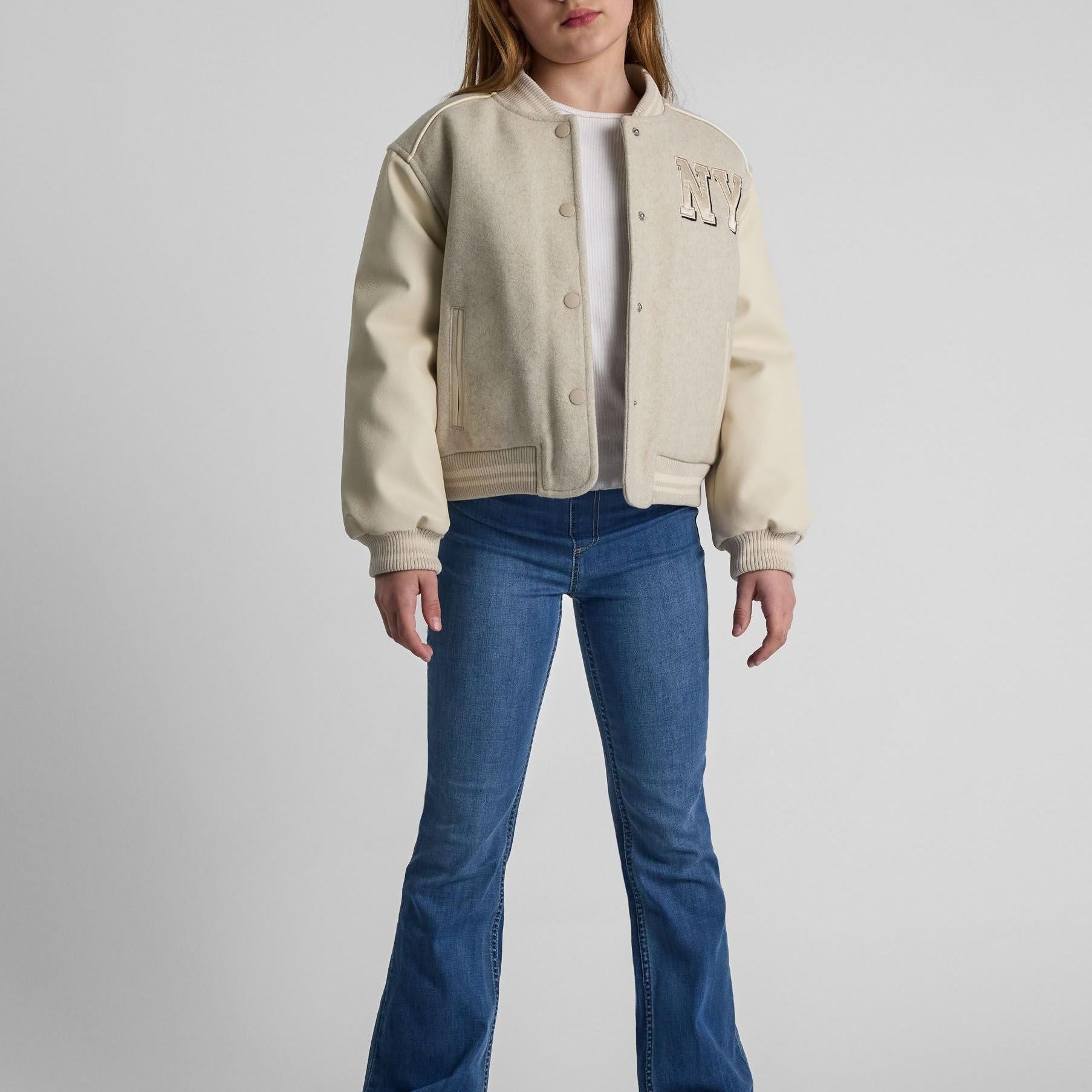 Girl wearing beige baseball jacket with faux leather sleeves and blue jeans against white background, front view showing NY patch detail and snap button closure
