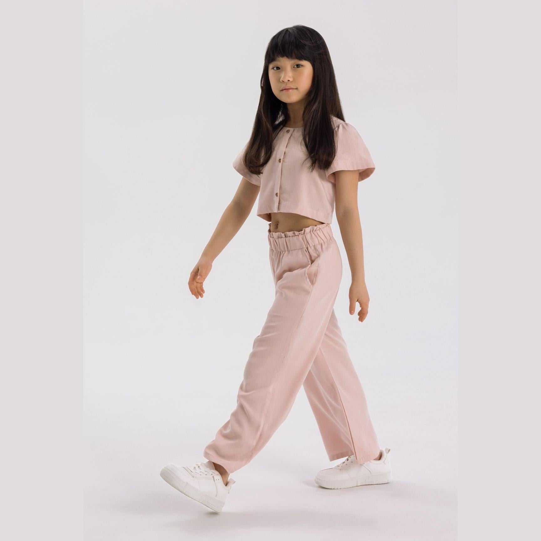 Young girl wearing pink blouse and wide leg trousers set, walking pose against white background