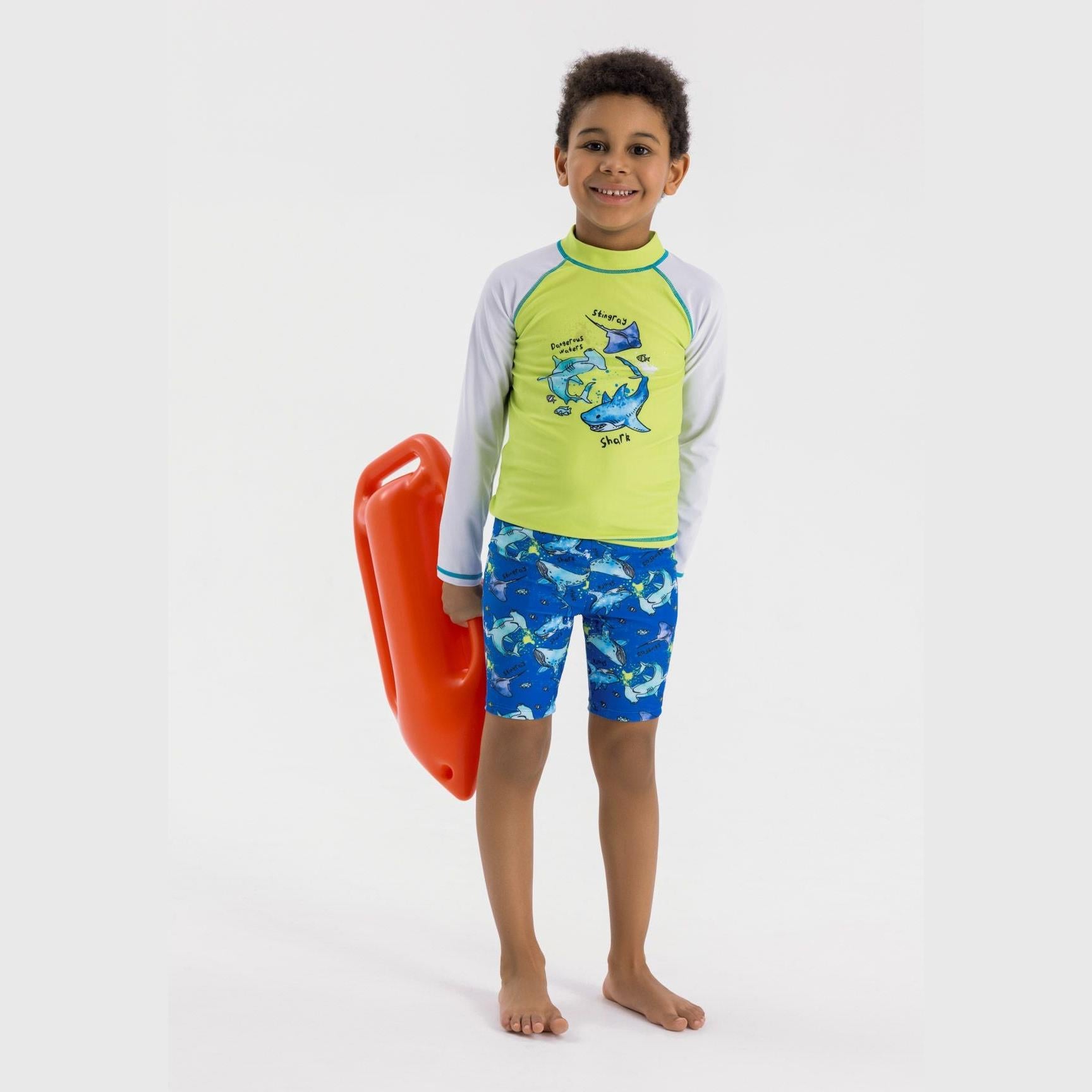 Young boy wearing green and white long sleeve swim top with shark graphics and blue patterned swim shorts, holding red bodyboard on white background