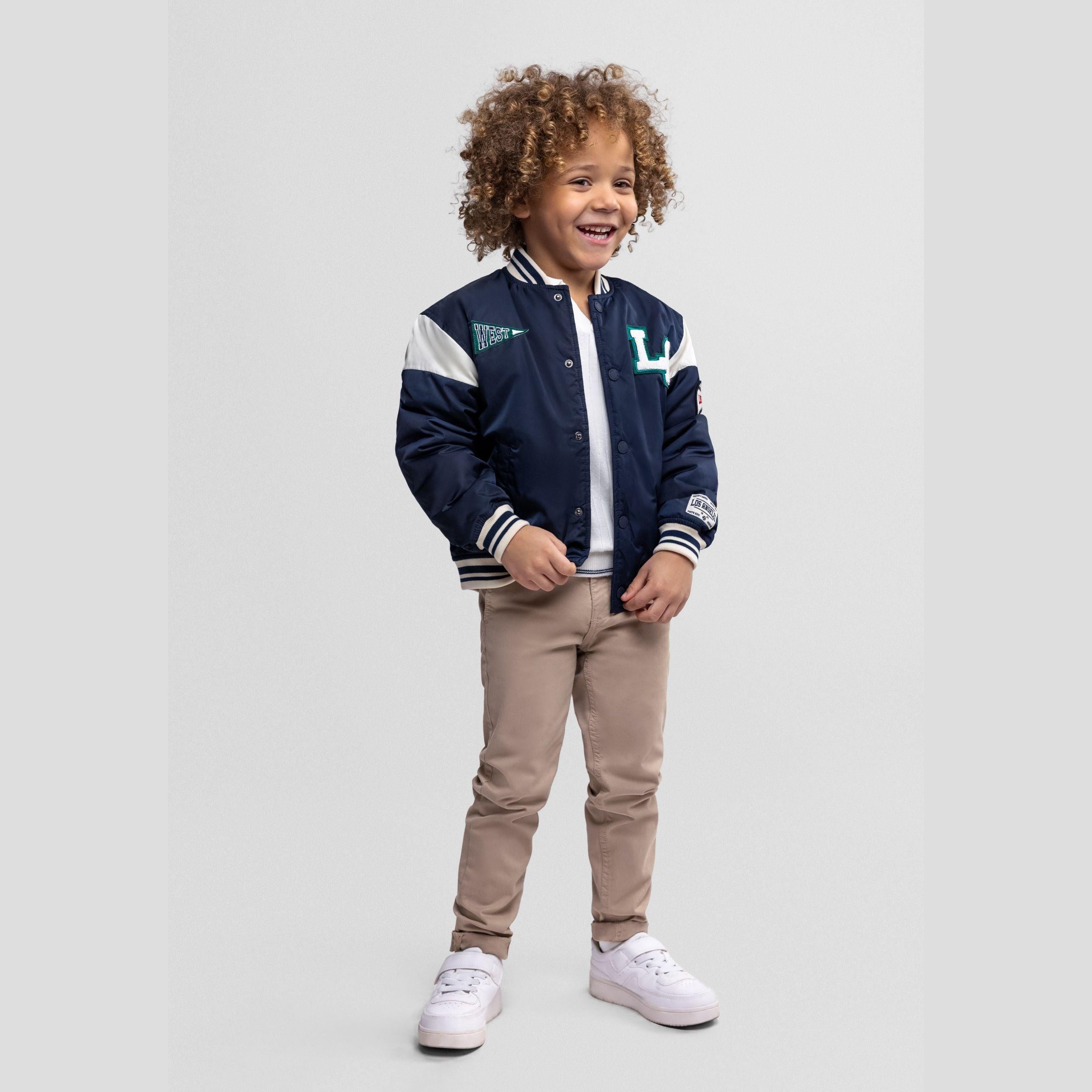 Young child with curly hair wearing navy varsity bomber jacket with white sleeves and beige trousers, smiling against white background