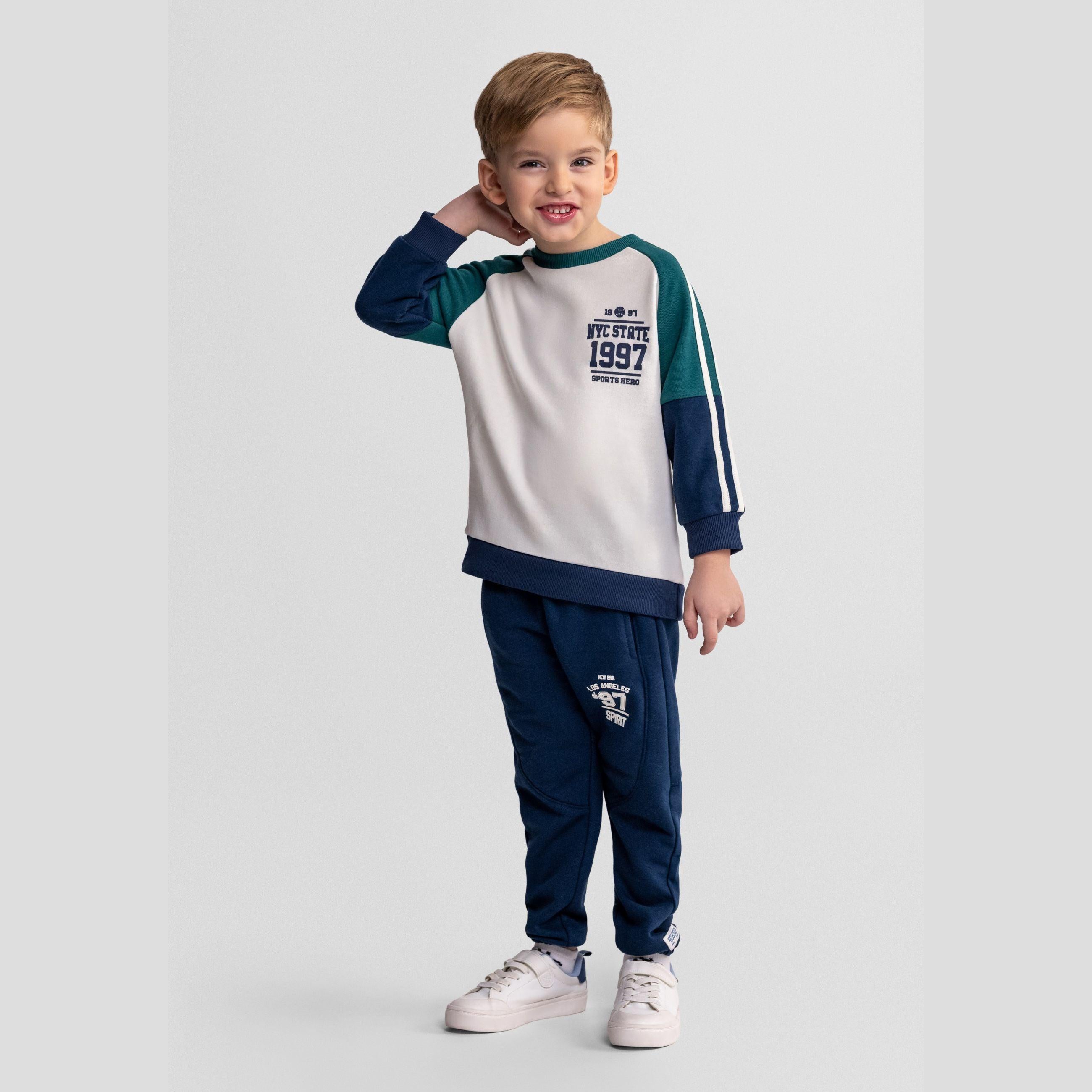 Young boy wearing navy tracksuit with green contrast panels posing playfully against light grey background, showing full outfit of sweatshirt and joggers