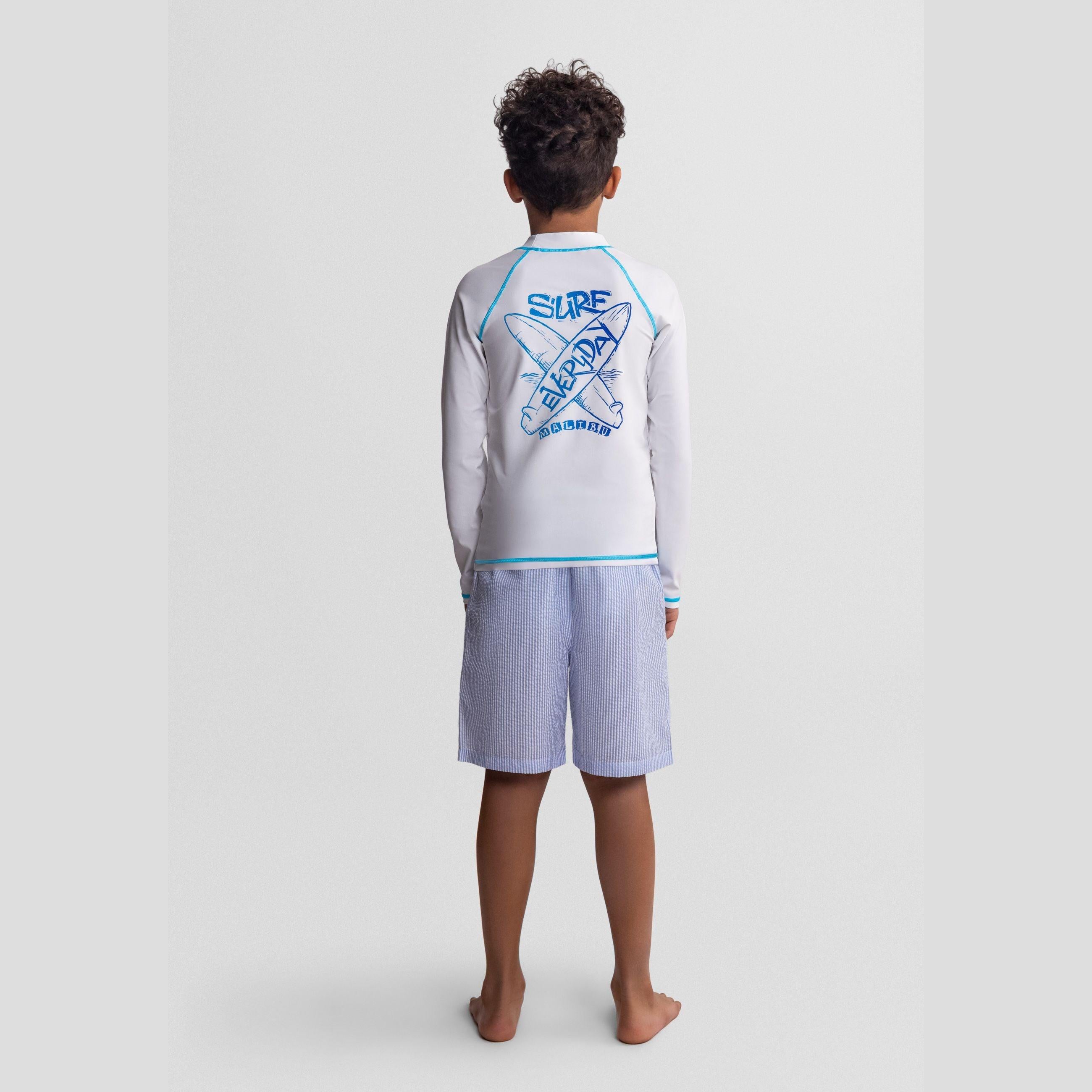 Back view of boy wearing white long sleeve rash vest with blue surf graphics and striped blue shorts against light background
