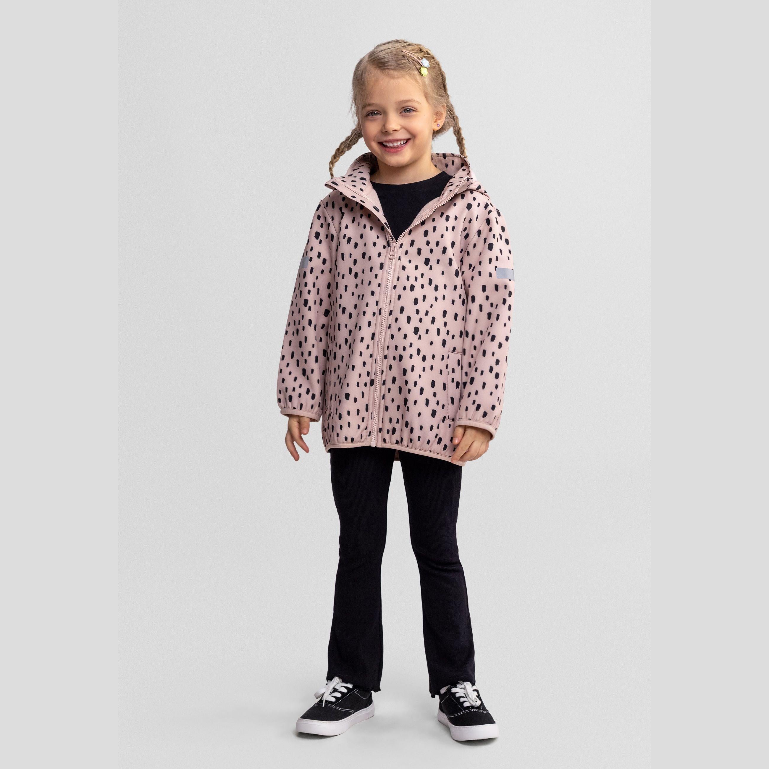 Young girl wearing pink softshell jacket with black spots over black outfit, standing against light grey background with blonde braided hair and yellow hair accessory