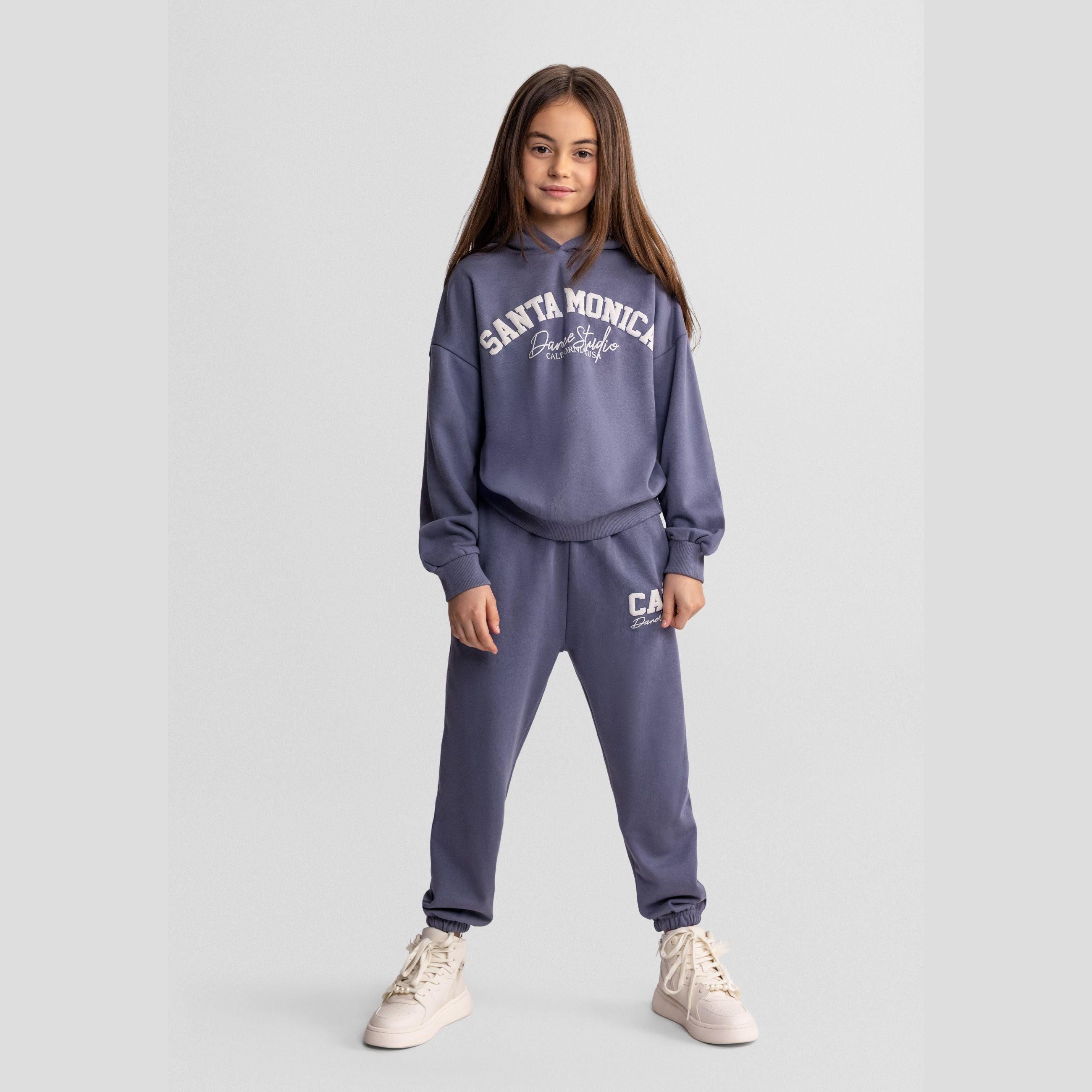 Young girl wearing blue-grey Santa Monica tracksuit with hooded sweatshirt and matching tapered joggers, standing against light grey background