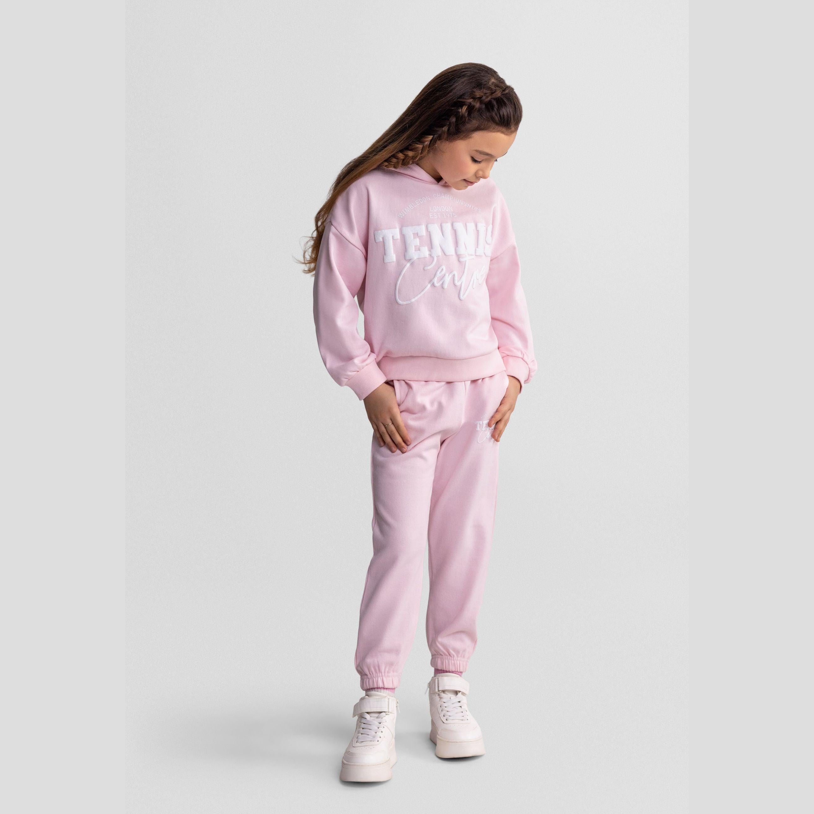 Girl wearing light pink tracksuit with Tennis Camps print, full body view against white background, casual standing pose
