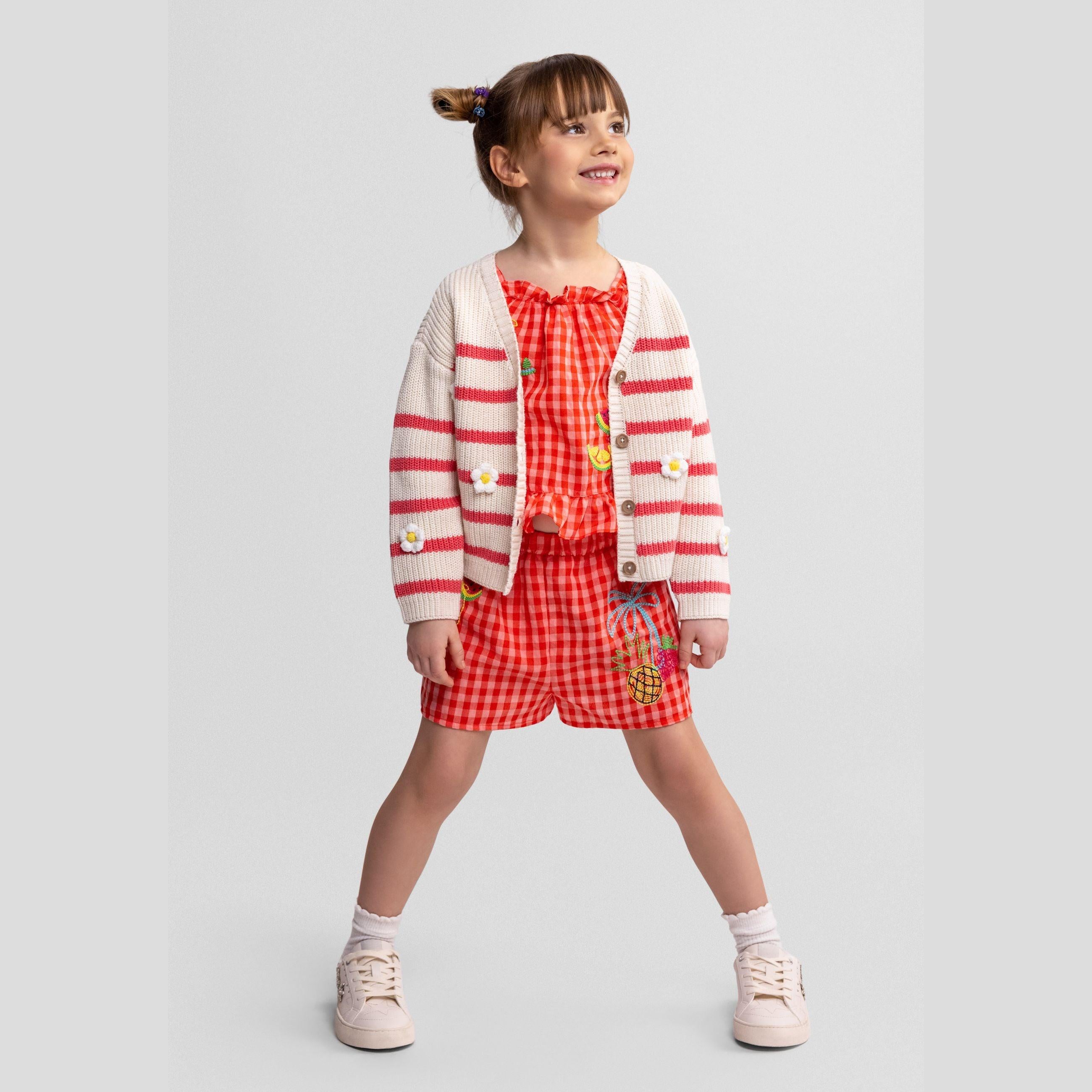 Girl wearing coral and white striped cardigan over red gingham dress, standing against light grey background