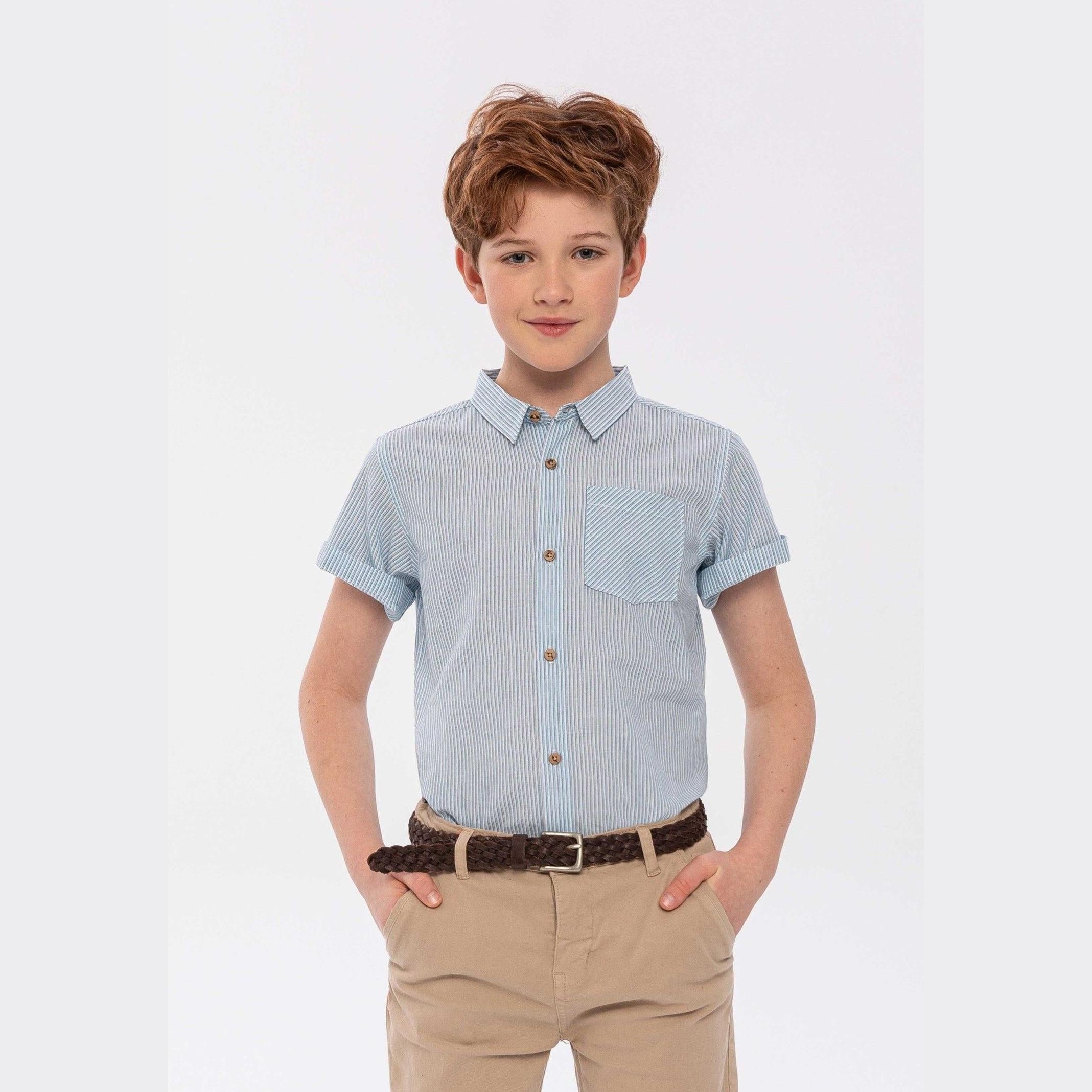 Boy wearing blue and white striped short sleeve poplin shirt with beige chinos and brown braided belt, standing against white background