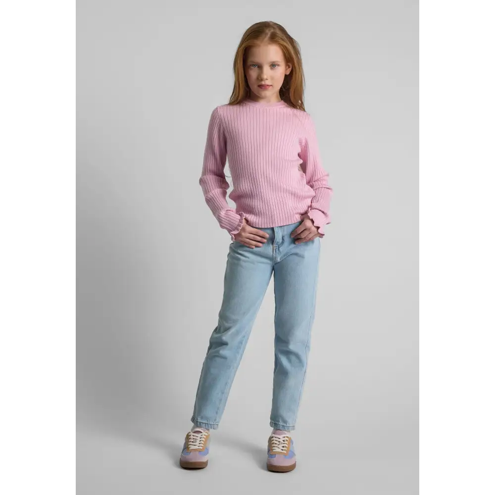 Young girl wearing light pink ribbed jumper with frill cuffs, paired with light blue jeans and pink sneakers, standing against white studio background