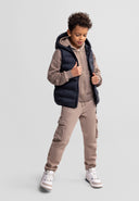 Boy wearing black quilted waistcoat with hood over beige hoodie and cargo pants in casual kids fashion style
