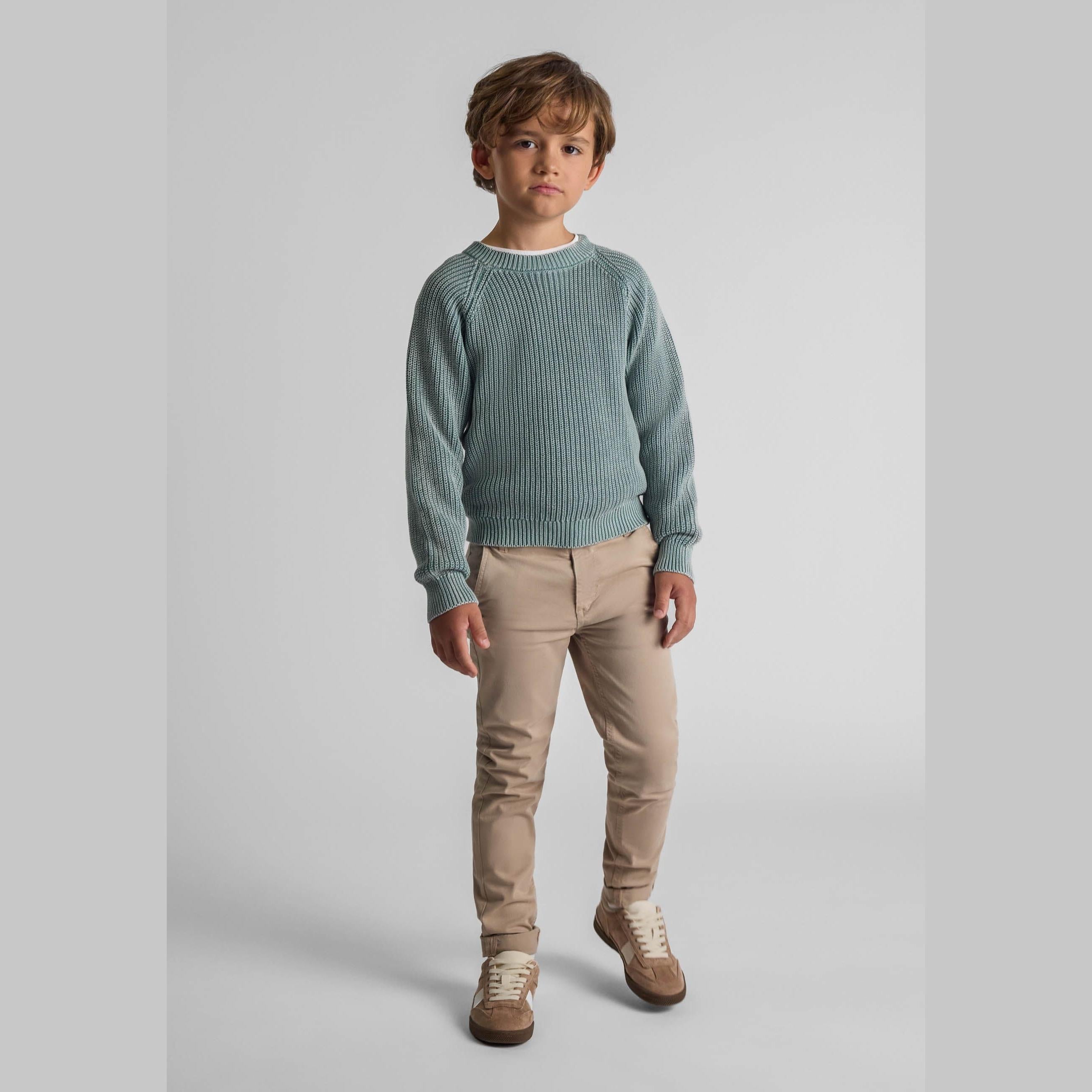 Young boy wearing khaki knit jumper with raglan sleeves and beige trousers, standing against light grey background in studio setting