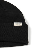Detail of a black ribbed beanie hat with a cuff and a logo badge.
