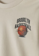 Detail of boys oversized cream sweatshirt with round neckline and basketball graphic.