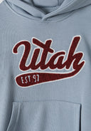 Detail of a blue boys oversized hoodie featuring a kangaroo pocket and embroidered 'Utah' logo.