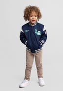 Toddler boy wearing navy padded varsity bomber jacket with striped collar and patches, paired with beige pants and white sneakers.