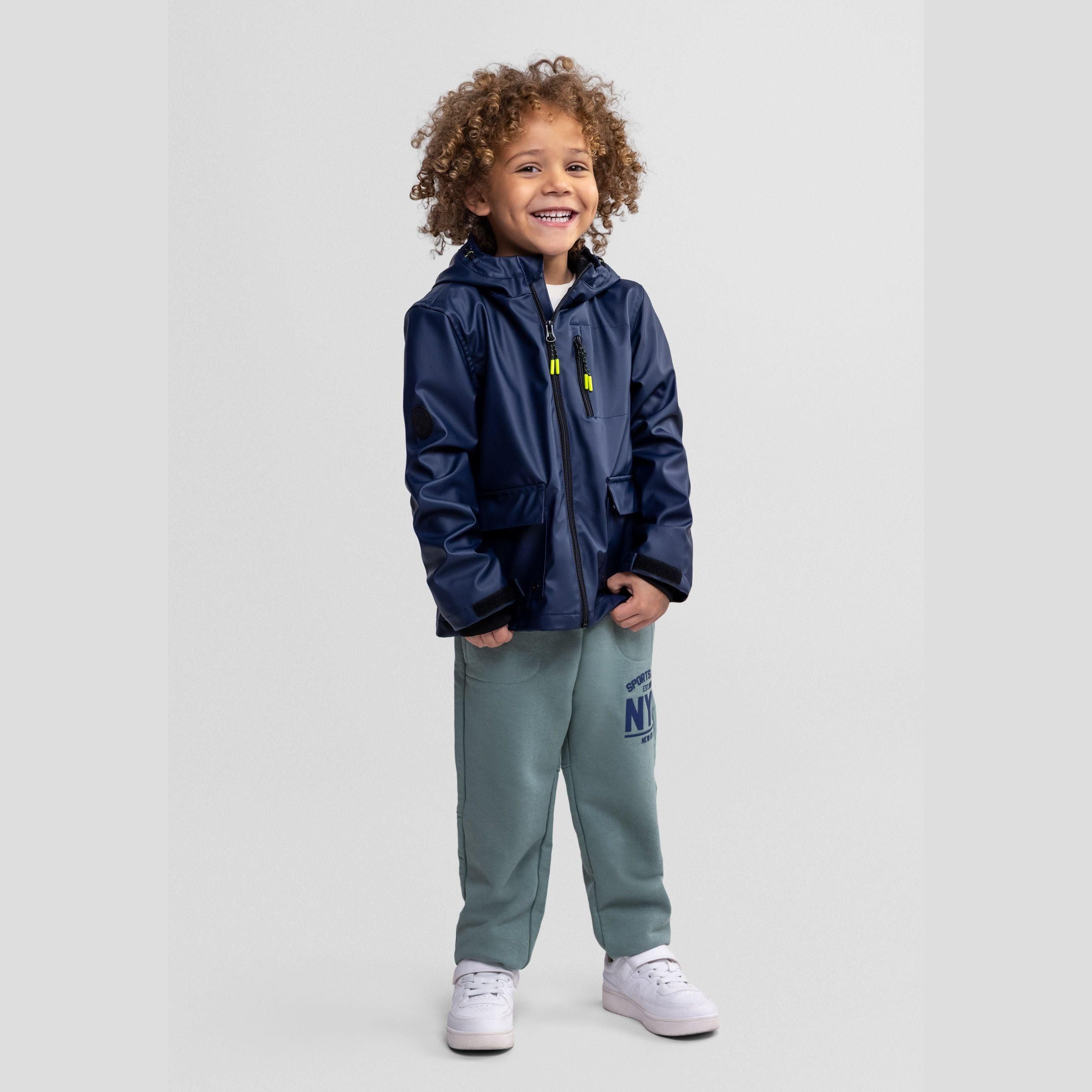 Smiling toddler wearing navy waterproof parka jacket with hood and sage green joggers against light grey background