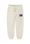 Cream-colored boys joggers with elastic waist, drawstring, side pockets, ribbed cuffs, and a black patch design.