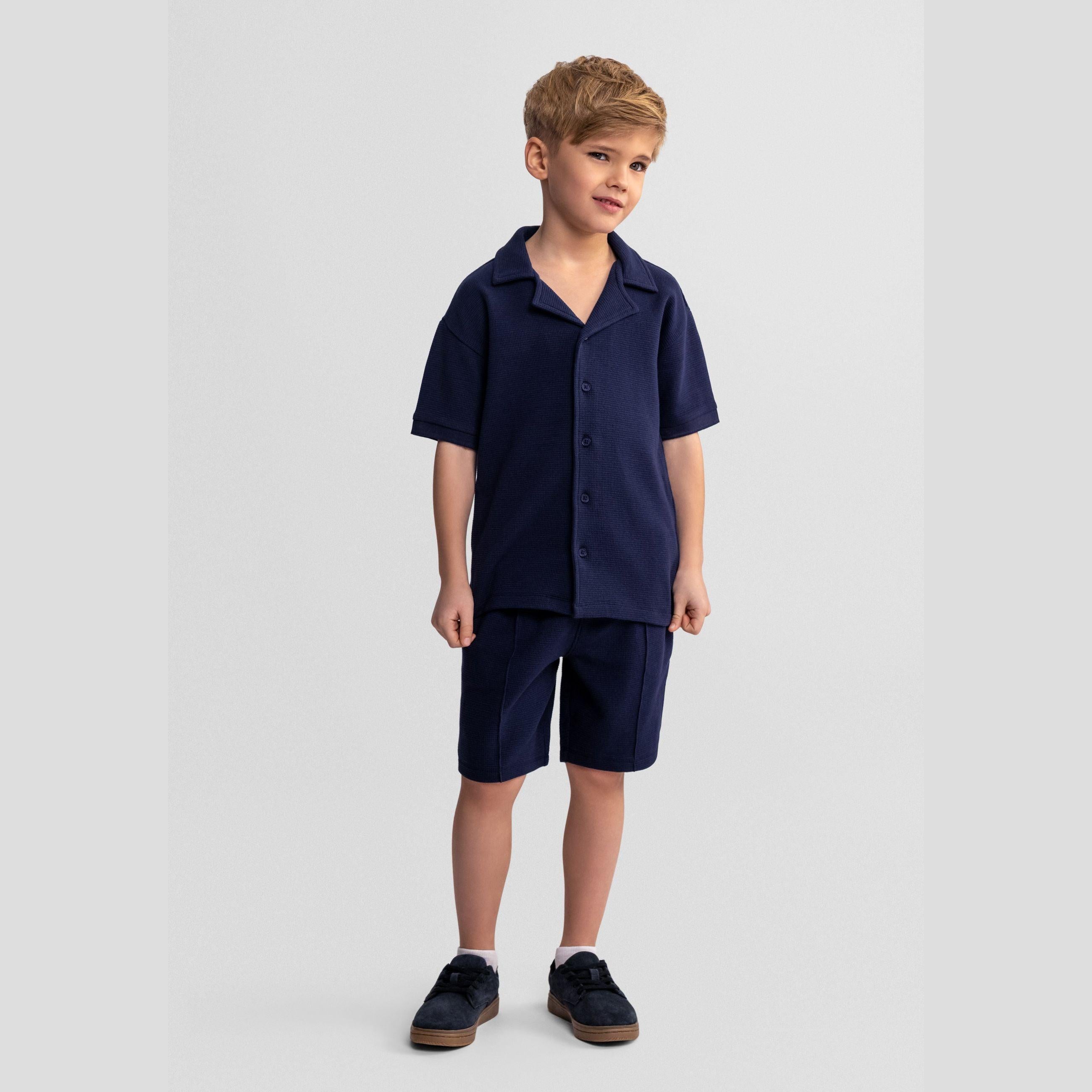 Young boy wearing navy blue polo shirt and shorts set, standing against light grey background with navy canvas shoes