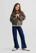Smiling girl wearing a stylish lightweight khaki bomber jacket with padded design and casual blue jeans standing confidently.
