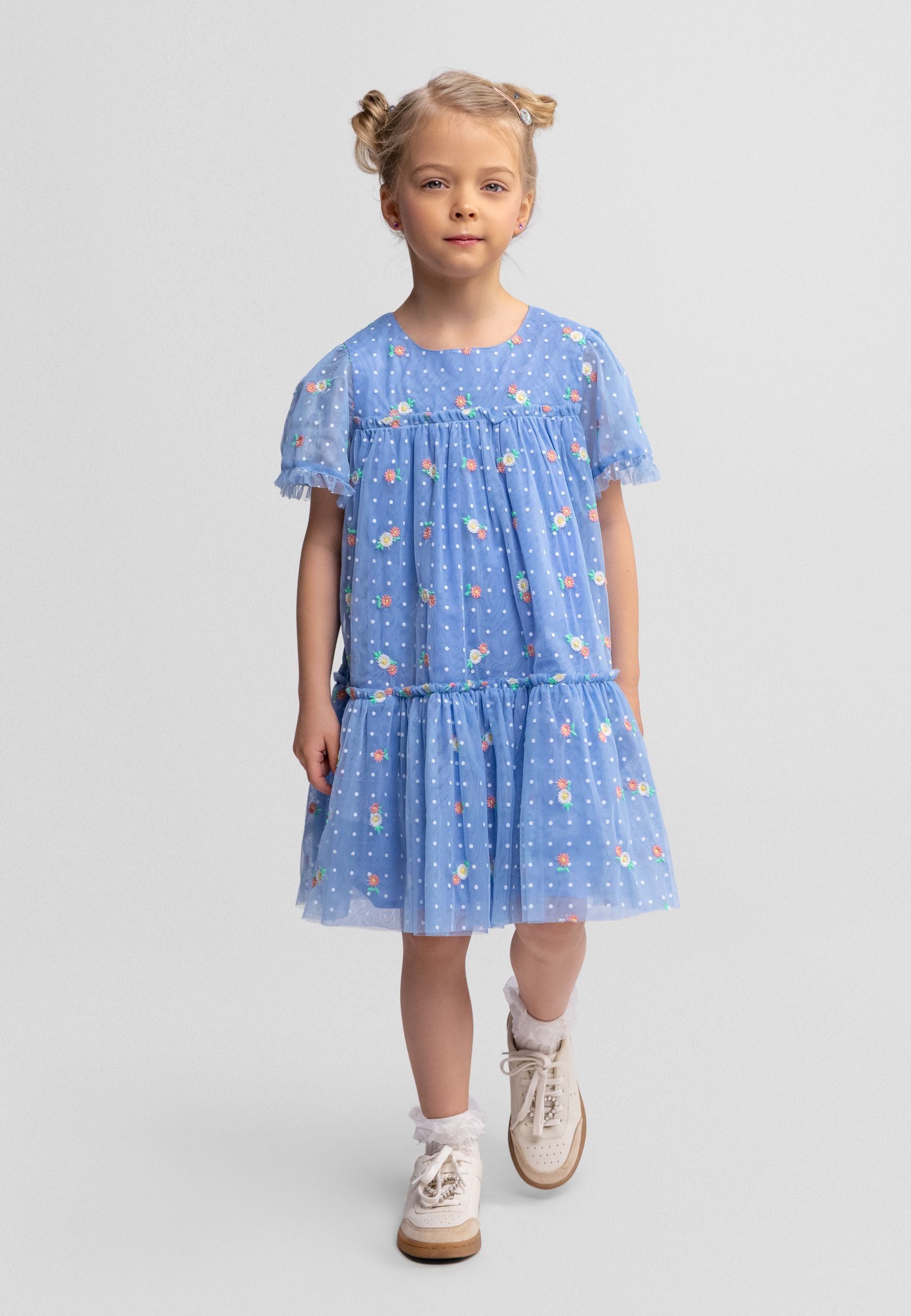 Young girl wearing blue tulle dress with floral embroidery and tiered skirt, walking against white background