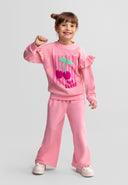Girl wearing a pink tracksuit with cherry design and ruffled sleeves, smiling in a playful pose against a plain background