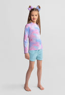 Young girl wearing a colorful pink and blue gradient long sleeve rash vest with light blue shorts standing barefoot.